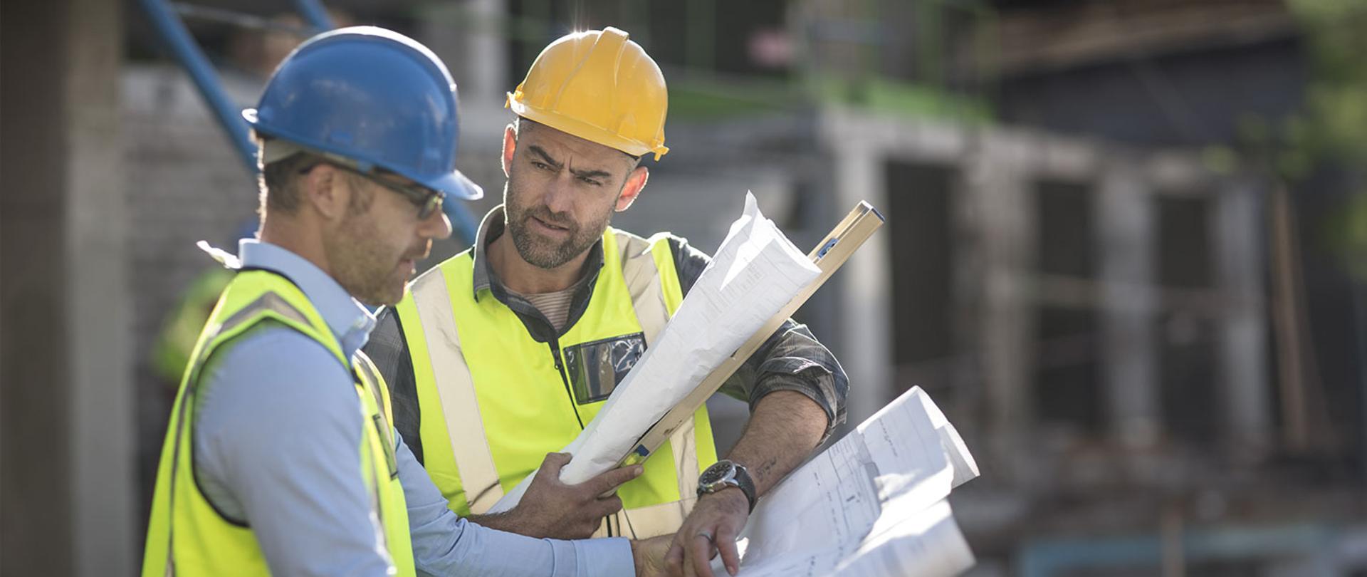 Zdjęcie przedstawia dwóch pracowników budowlanych w kaskach i kamizelkach odblaskowych z planami w dłoniach.