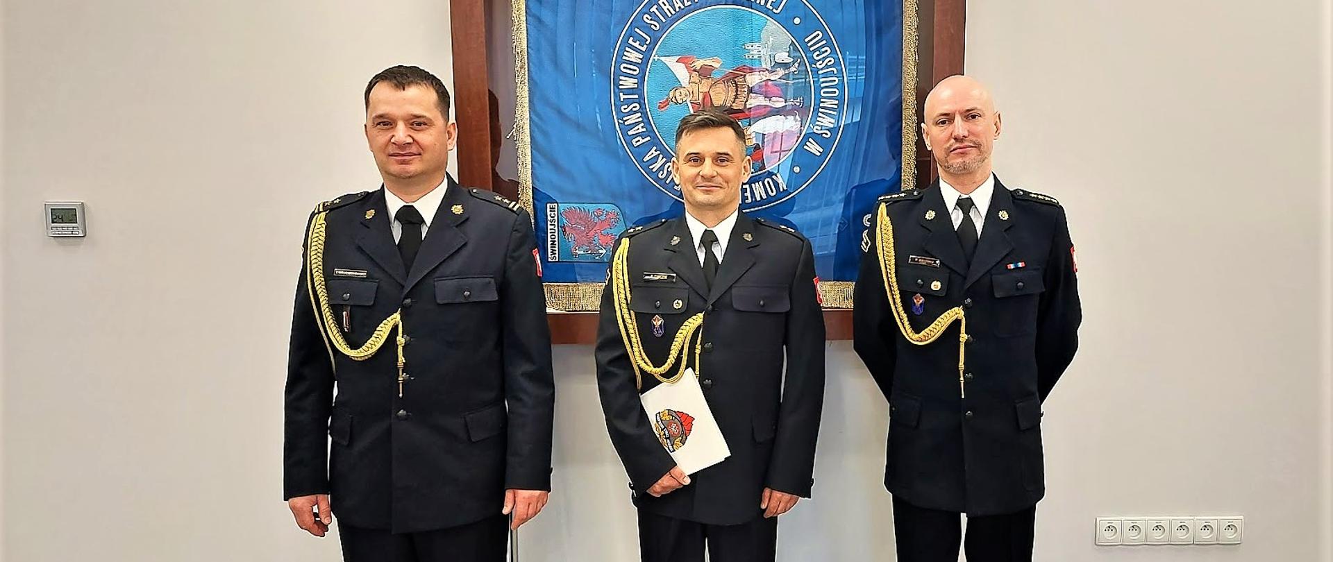 3 oficerowie PSP w mundurach wyjściowych ze sznurem en face na tle sztandaru komendy. 