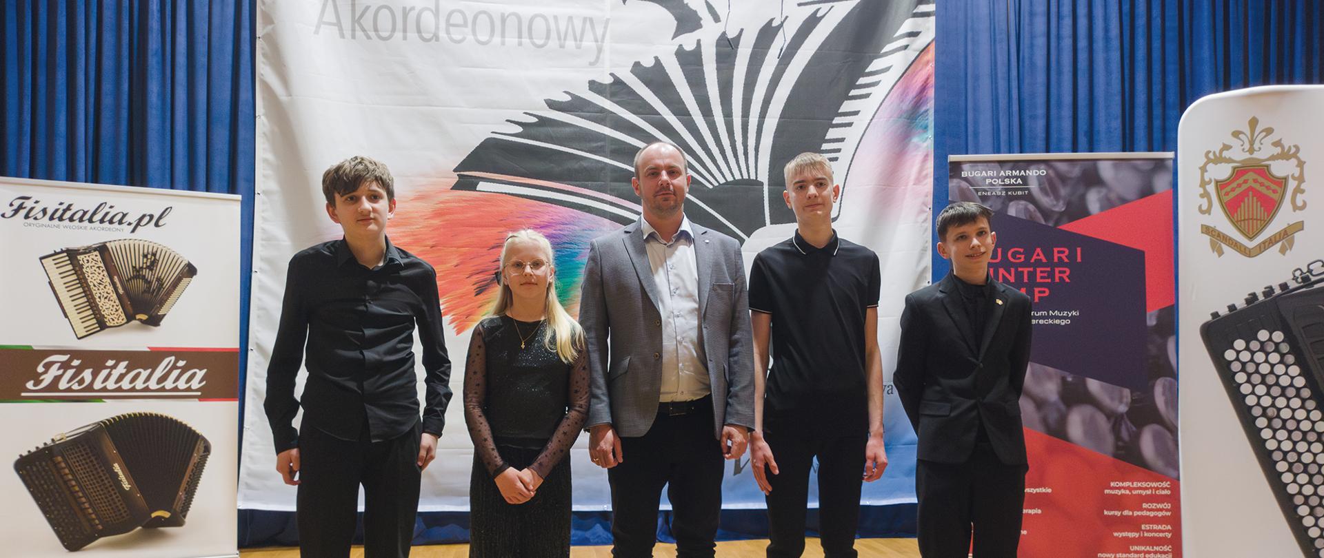 Ogólnopolski konkurs akordeonowy – pięć osób (czterech chłopców i jedna dziewczynka) oraz dorosły mężczyzna stoją na scenie przed dużym banerem z grafiką akordeonu; uczestnicy ubrani na czarno, w tle roll-upy sponsorów, na podłodze bukiet niebieskich kwiatów.