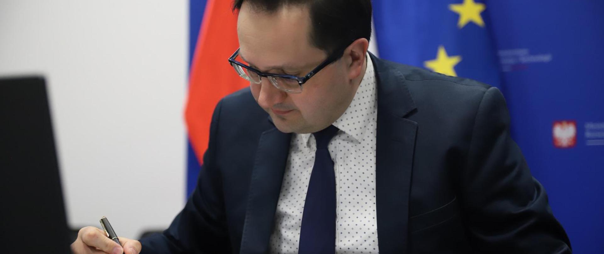 Wiceminister przy stole z piórem w reku podpisuje dokument