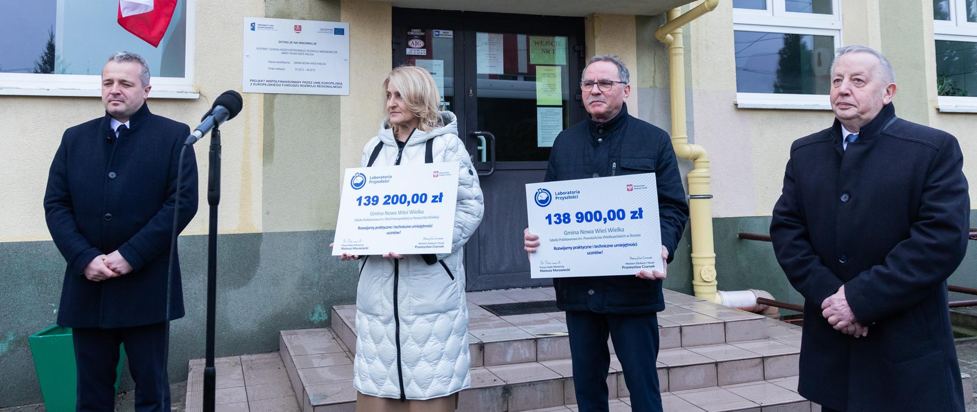 Trzech mężczyzn i jedna kobieta stojący przed budynkiem. Dwoje spośród nich trzyma vouchery. 