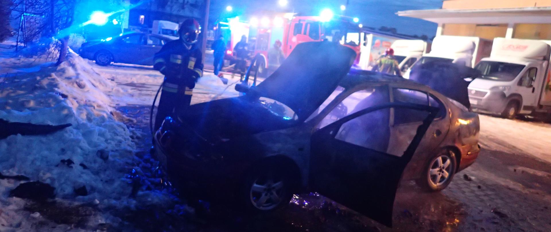 Na zdjęciu widoczny jest pojazd, który jest gaszony prądem wody przez strażaka. W tle znajdują się pojazdy firmowe oraz zastęp PSP.