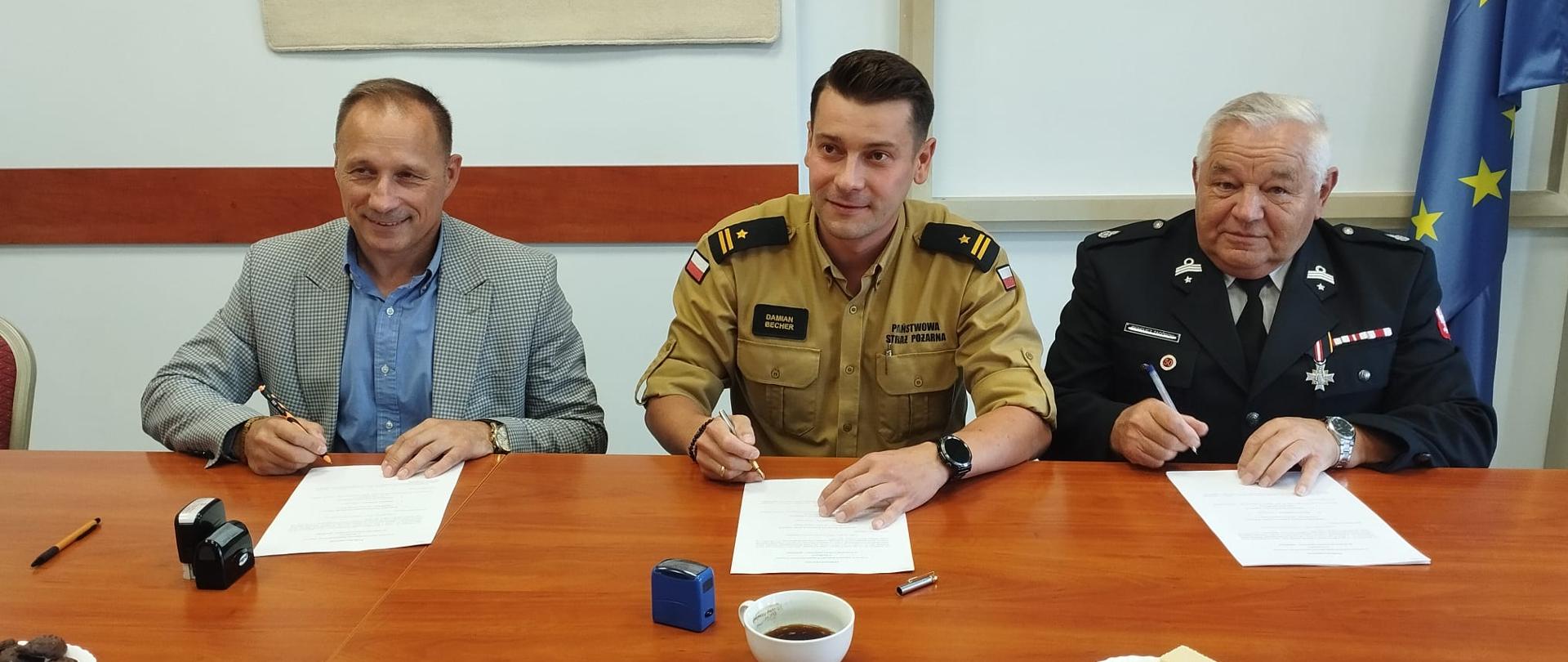 Zdjęcie przedstawia 3 mężczyzn, jeden w ubraniu strażackim służbowym, drugi w mundurze OSP, a trzeci w garniturze. Siedzą przy stole i podpisują dokumenty. Za nimi na ścianie widnieje fragment godła państwowego oraz fragment sztandaru.