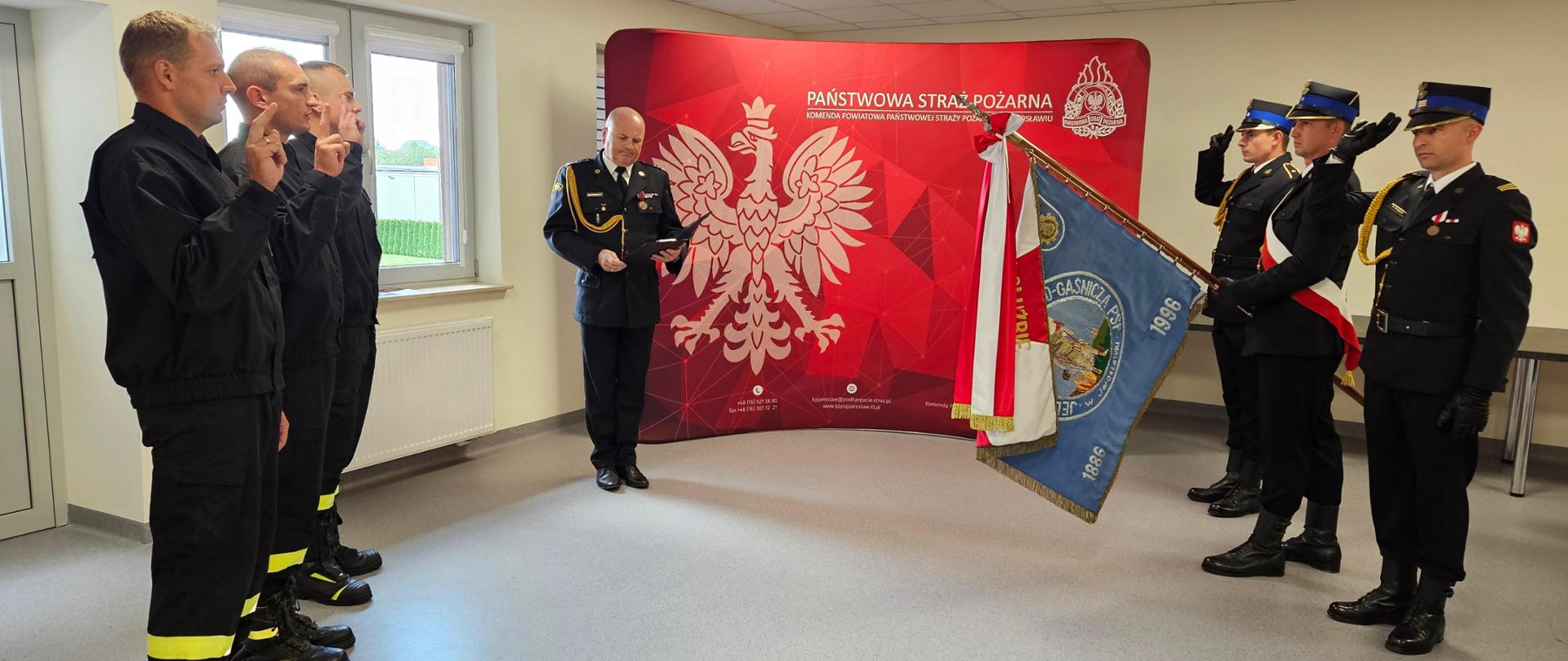 Zdjęcie wykonane w sali szkoleniowej Komendy Powiatowej Państwowej Straży Pożarnej w Jarosławiu w trakcie składania ślubowania przez nowych funkcjonariuszy. Po lewej stoi 3 funkcjonariuszy w postawie jak do ślubowania, po prawej salutuje poczet sztandarowy, a w centralnej części stoi Komendant Powiatowy, który odczytuje słowa ślubowania. W tle widoczna jest ścianka promocyjna.