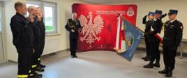 Zdjęcie wykonane w sali szkoleniowej Komendy Powiatowej Państwowej Straży Pożarnej w Jarosławiu w trakcie składania ślubowania przez nowych funkcjonariuszy. Po lewej stoi 3 funkcjonariuszy w postawie jak do ślubowania, po prawej salutuje poczet sztandarowy, a w centralnej części stoi Komendant Powiatowy, który odczytuje słowa ślubowania. W tle widoczna jest ścianka promocyjna.