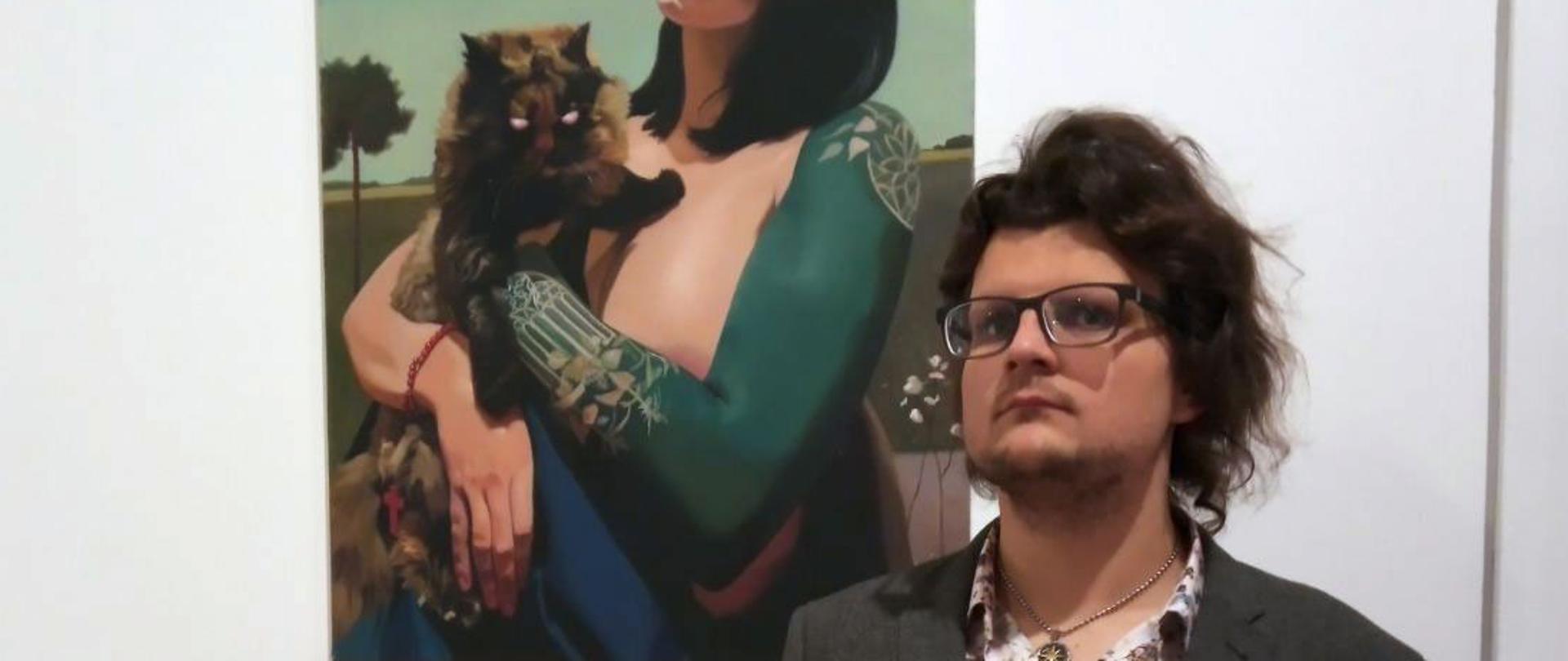 Na pierwszym planie Michał Rostkowski, w tle jego obraz przedstawiający kobietę trzymającą kota na ręku.