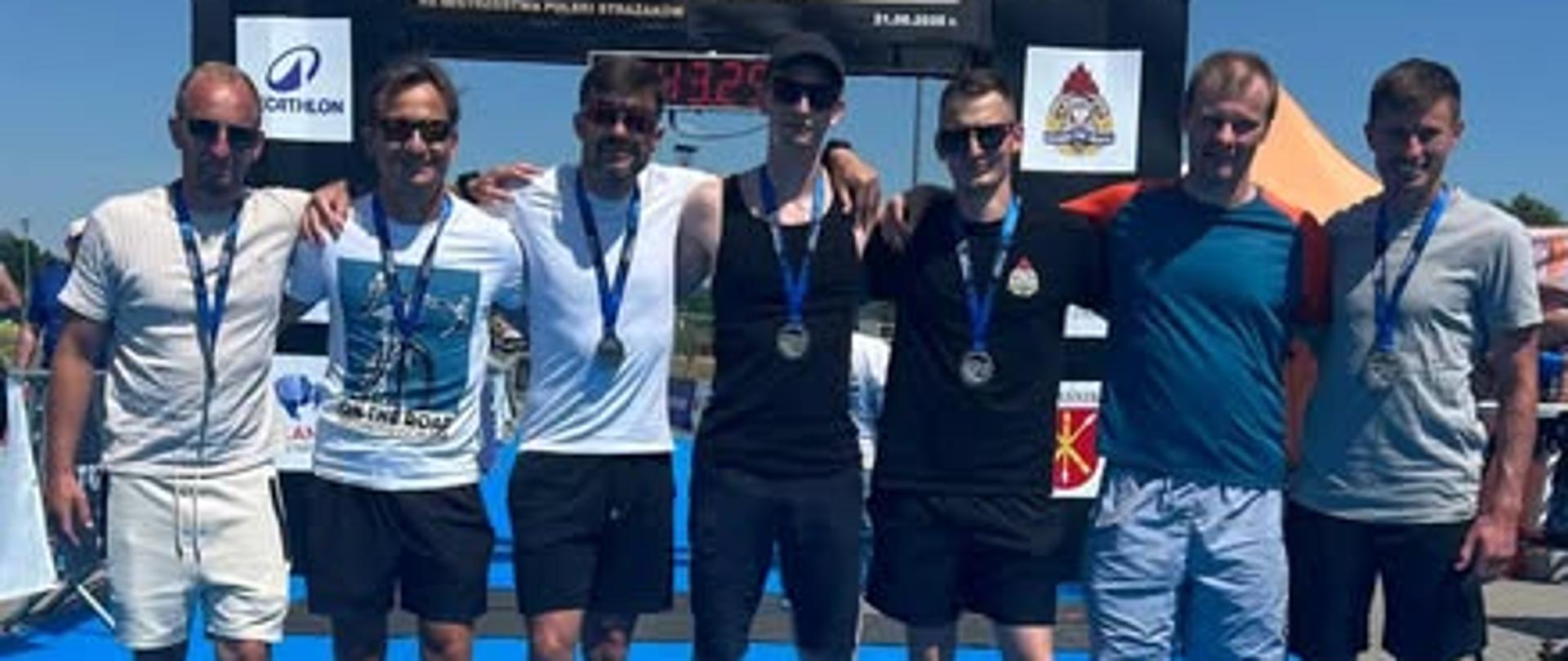 Na zdjęciu widzimy grupę siedmiu mężczyzn stojących ramię w ramię na jasnoniebieskim dywanie, prawdopodobnie tuż po zakończeniu zawodów sportowych.
Oto szczegóły, które można zauważyć:
Tło i sceneria: Mężczyźni pozują na tle dużej czarnej bramy lub tablicy z napisem „TRIATHLON KRAŚNIK”. Na tablicy widoczne są także mniejsze logotypy (m.in. marki Decathlon) oraz data wskazująca na rok 2020. Pogoda jest słoneczna, a niebo bezchmurne.
Uczestnicy: Większość mężczyzn ma na szyi zawieszone medale, co sugeruje, że ukończyli oni wspomniane zawody. Są ubrani w luźne, sportowe stroje – głównie t-shirty, koszulki bez rękawów oraz krótkie spodenki. Wielu z nich ma na sobie okulary przeciwsłoneczne, a na stopach klapki lub są boso.
Atmosfera: Wszyscy wyglądają na zadowolonych, uśmiechają się i obejmują, co wskazuje na udany start i przyjacielską, drużynową atmosferę.