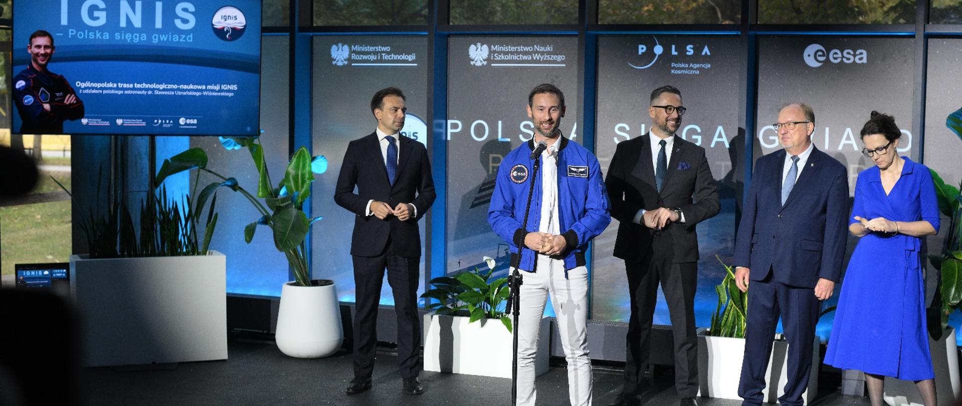 Na sali przed banerami z napisem "Polska sięga gwiazd" stoi pięć osób, drugi od lewej Sławosz Uznański-Wiśniewski mówi do mikrofonu na stojaku, z boku z lewej na dużym monitorze umieszczonym na słupie napis IGNIS, w tle duże okna.