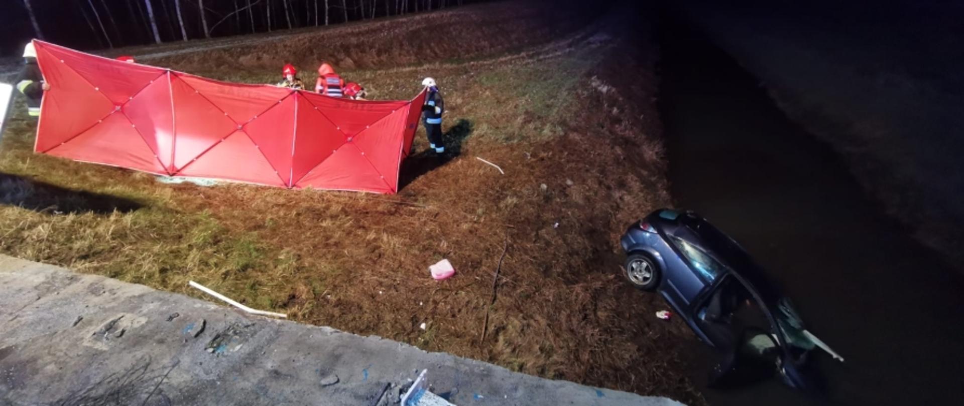 Zdjęcie zrobione o zmroku. Po prawej stronie fotografii widoczny samochód osobowy koloru granatowego częściowo zanurzony w kanale z wodą. Po lewej stronie zdjęcia rozstawiony czerwony parawan, za którym widać strażaków i personel ZRM.