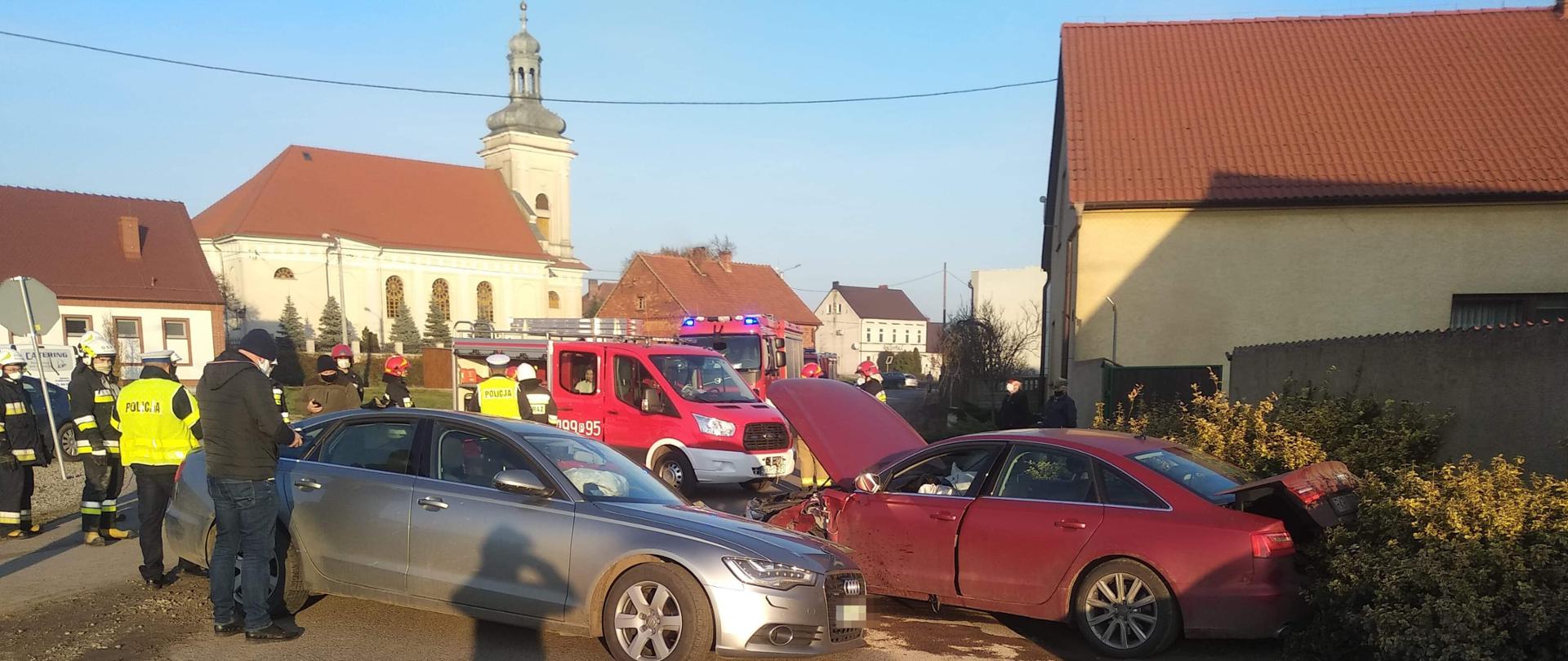 Uszkodzone środki transportu blokują drogę. Funkcjonariusze Policji prowadzą czynności na miejscu wypadku. W tle samochody pożarnicze oraz strażacy w ubraniach bojowych. Na jezdni widoczne wycieki płynów eksploatacyjnych. Samochody uczestniczące w zdarzeniu mają rozbite przody. 