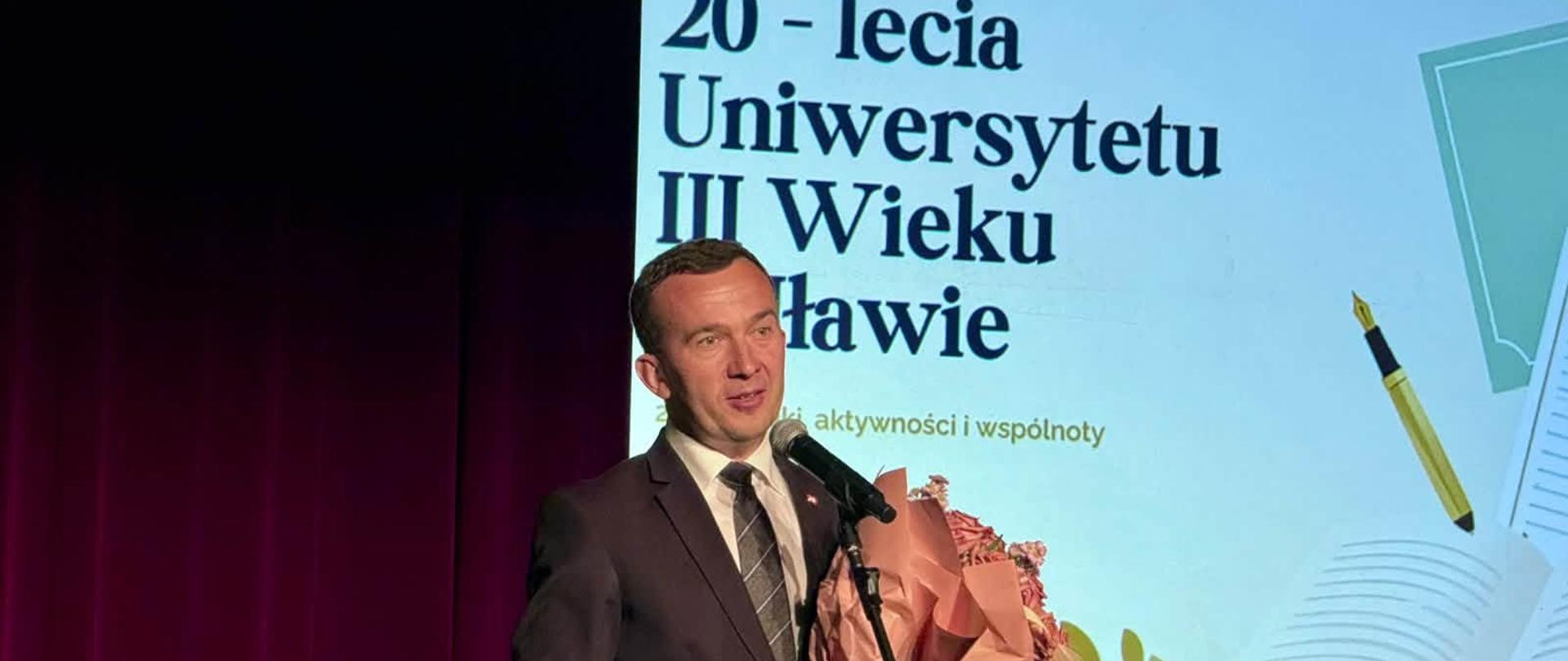 Mężczyzna w garniturze wygłasza przemówienie podczas jubileuszu Uniwersytetu Trzeciego Wieku w Iławie