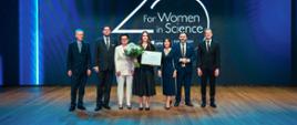 Na tle granatowej ściany z napisem 25 for Women in Science stoi czterech mężczyzn w garniturach i trzy kobiety, drugi od lewej wiceminister Szeptycki, kobieta pośrodku trzyma dokument w ramkach i bukiet białych kwiatów.