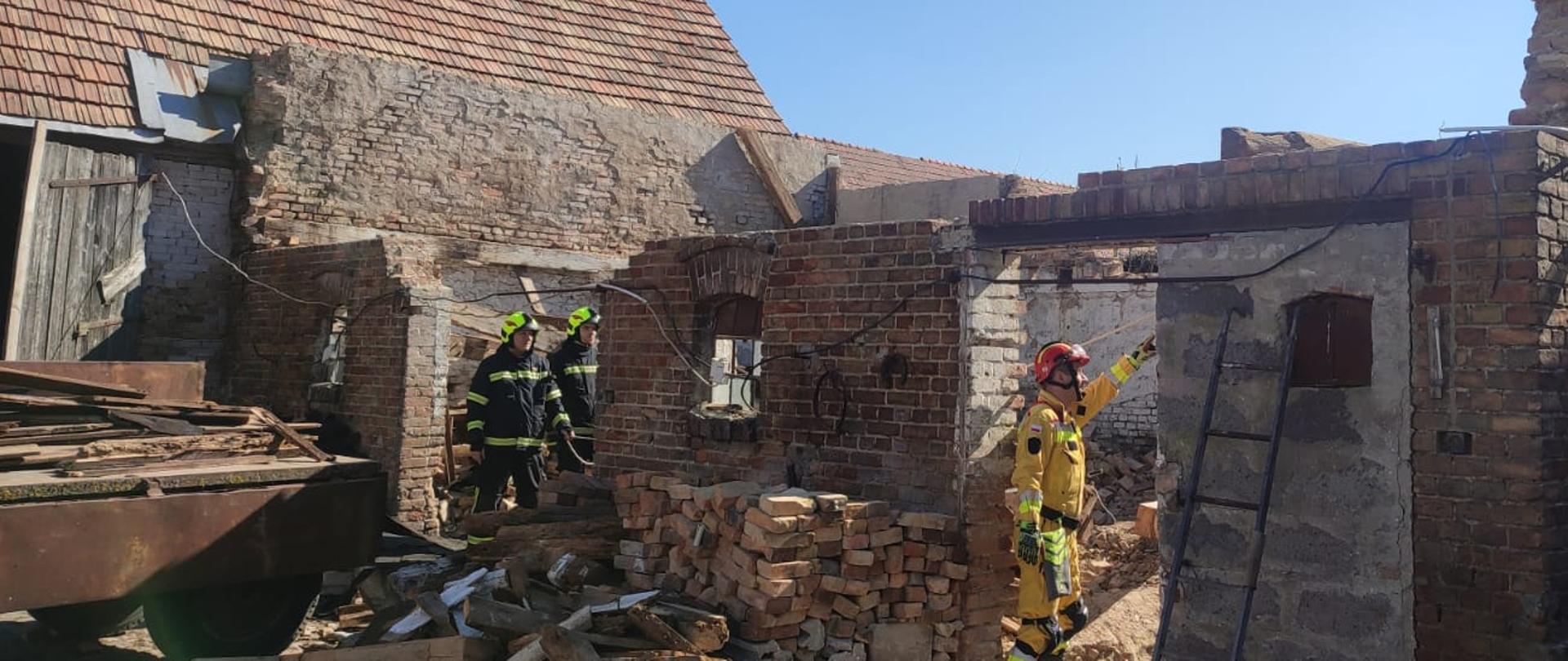 Na zdjęciu ceglany budynek bez dachu, w otworach po drzwiach stoją strażacy. Po prawej stronie widać ratownika medycznego.