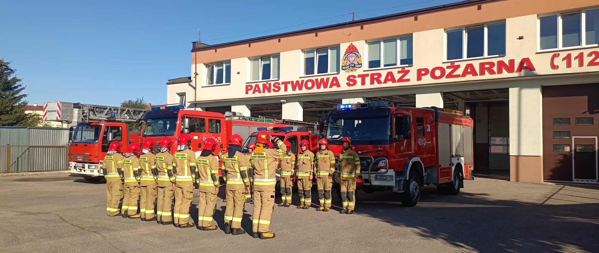 Fotografia przedstawia strażaków z JRG Malbork oddających hołd minutą ciszy strażakowi, który zginął w wypadku podczas służby. Zmiany służbowe stoją w ubraniach bojowych na przeciwko siebie na baczność. W tle widać pojazdy pożarnicze z włączonymi sygnałami świetlnymi koloru niebieskiego oraz budynek Komendy Powiatowej Państwowej Straży Pożarnej.