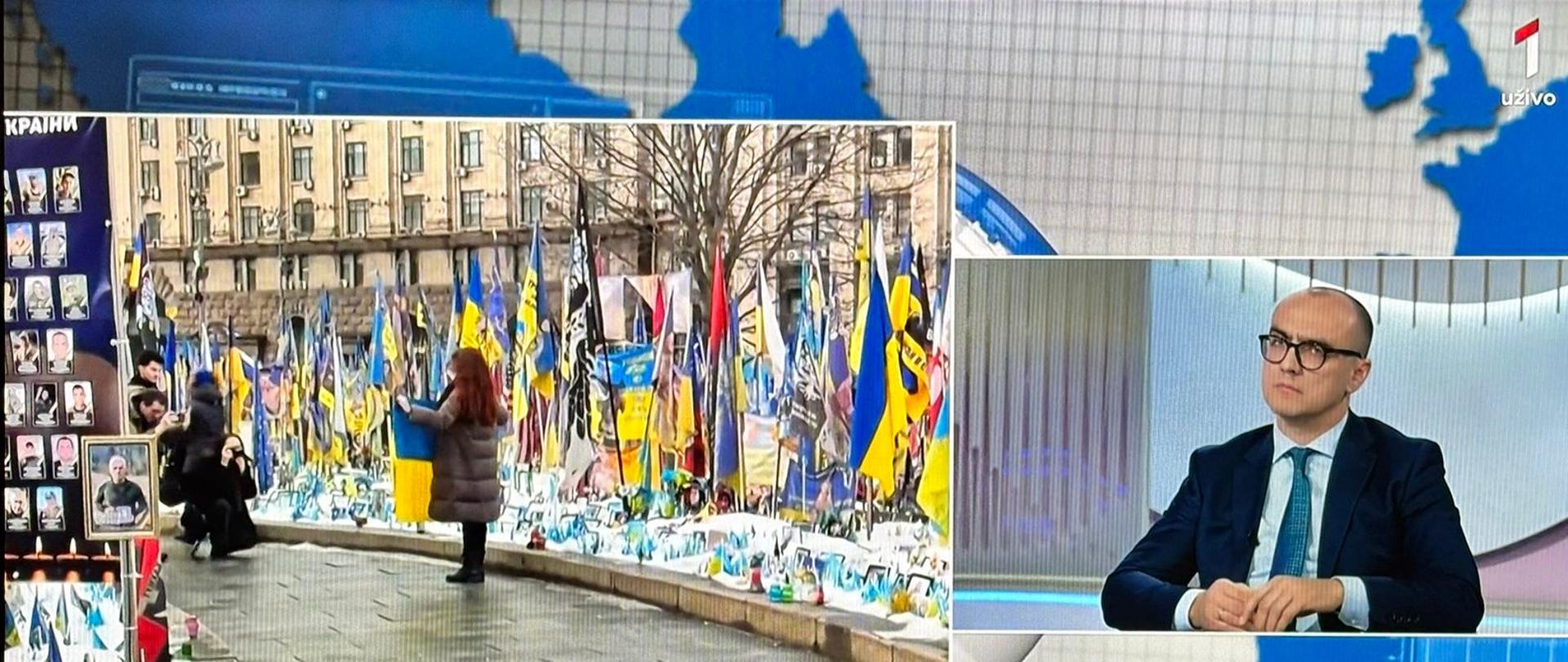 Chargé d'affaires a.i. Bartosz Marcinkowski w programie Globus o polskim wsparciu dla Ukrainy_Obok zdjęcie z flagami Ukrainy, zniczami i zdjęciami ofiar wojny.