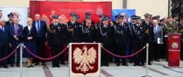Za godłem Polski stoją strażacy ubrani w mundury galowe oraz zaproszenie goście z tyłu widać budynek ratusza