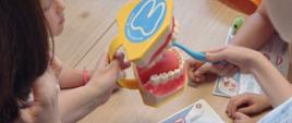 Kobieta korzystając z modelu szczęki i szczotki pokazuje dzieciom jak prawidłowo szczotkować zęby. 
