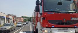 Słoneczny dzień, ruchliwa ulica miasta. Na pasie w przeciwnym kierunku ruchu zaparkowany czerwony samochód strażacki z włączonymi światłami mijania oraz ostrzegawczymi koloru niebieskiego. W kabinie strażackiej ciężarówki widać sylwetkę człowieka. Na pasie przeciwnym widać sznur pojazdów osobowych. 