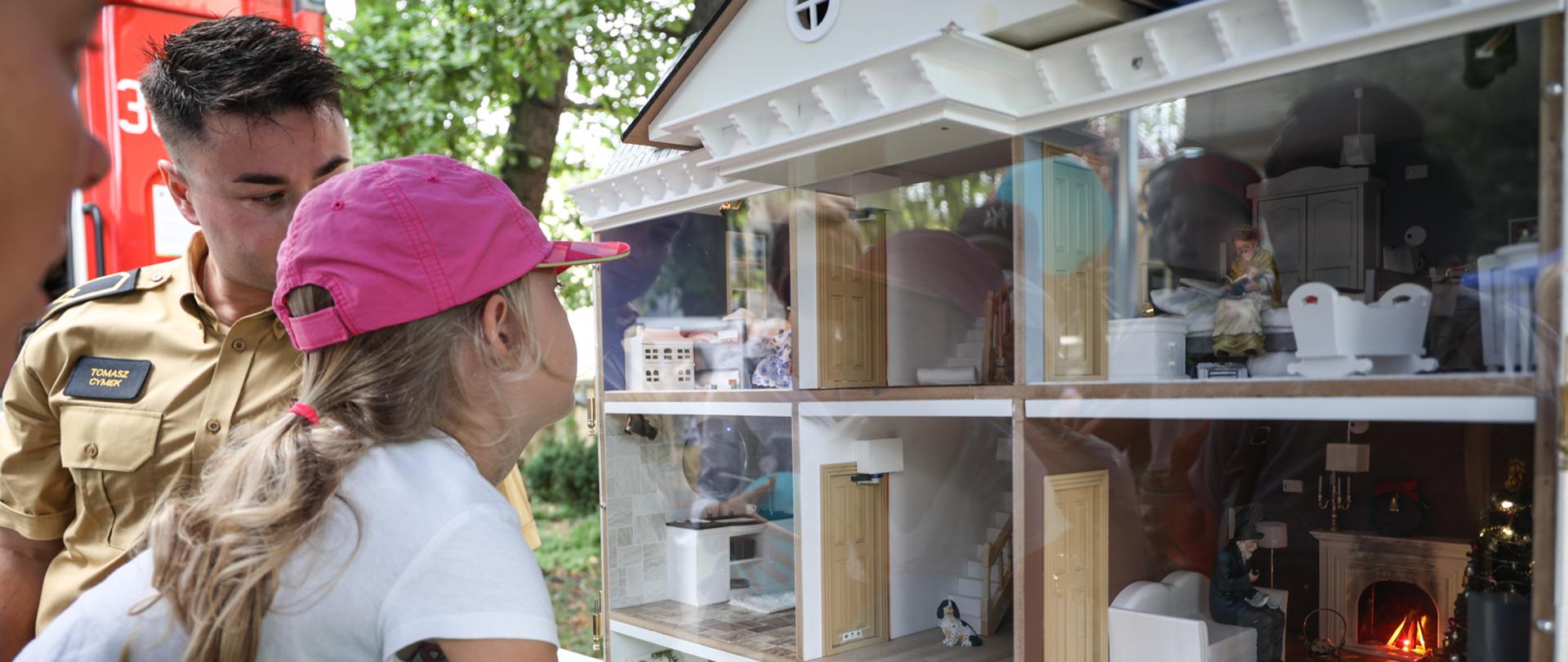 Strażak ubrany w mundur koloru musztardowego przedstawia dziewczynce w różowej czapce z daszkiem domek opisujący zagrożenia które mogą wystąpić w mieszkaniu lub domu. 