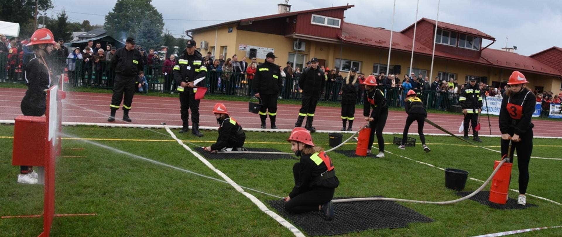 Kolorowa fotografia wykonana w pochmurny dzień na stadionie sportowym. Na pierwszy planie zawodniczki Młodzieżowej Drużyny Pożarniczej podczas nalewania hydronetkami wody do nalewaków. W głębi widoczne inne zawodniczki i kliku strażaków oceniających zadanie. W tle liczni zgromadzeni widzowie przy stadionie a za nimi duży budynek stadionowego zaplecza.