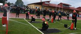 Kolorowa fotografia wykonana w pochmurny dzień na stadionie sportowym. Na pierwszy planie zawodniczki Młodzieżowej Drużyny Pożarniczej podczas nalewania hydronetkami wody do nalewaków. W głębi widoczne inne zawodniczki i kliku strażaków oceniających zadanie. W tle liczni zgromadzeni widzowie przy stadionie a za nimi duży budynek stadionowego zaplecza.