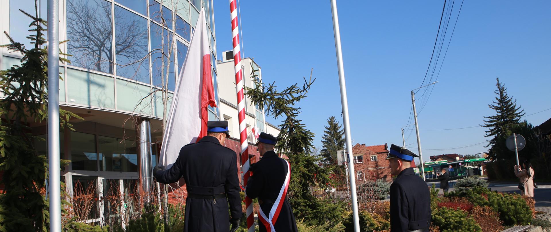 Trzech strażaków w mundurach wyjściowych zakłada flagę państwową na maszt. Uroczystość związana jest z Narodowym Dniem Niepodległości i odśpiewaniem hymnu państwowego. Na drugim planie budynek strażnicy.