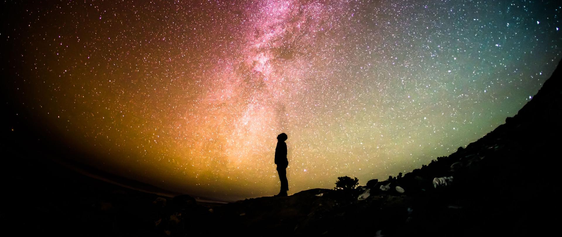 rozgwieżdżone niebo. Po środku ciemna sylwetka człowieka patrzącego do góry