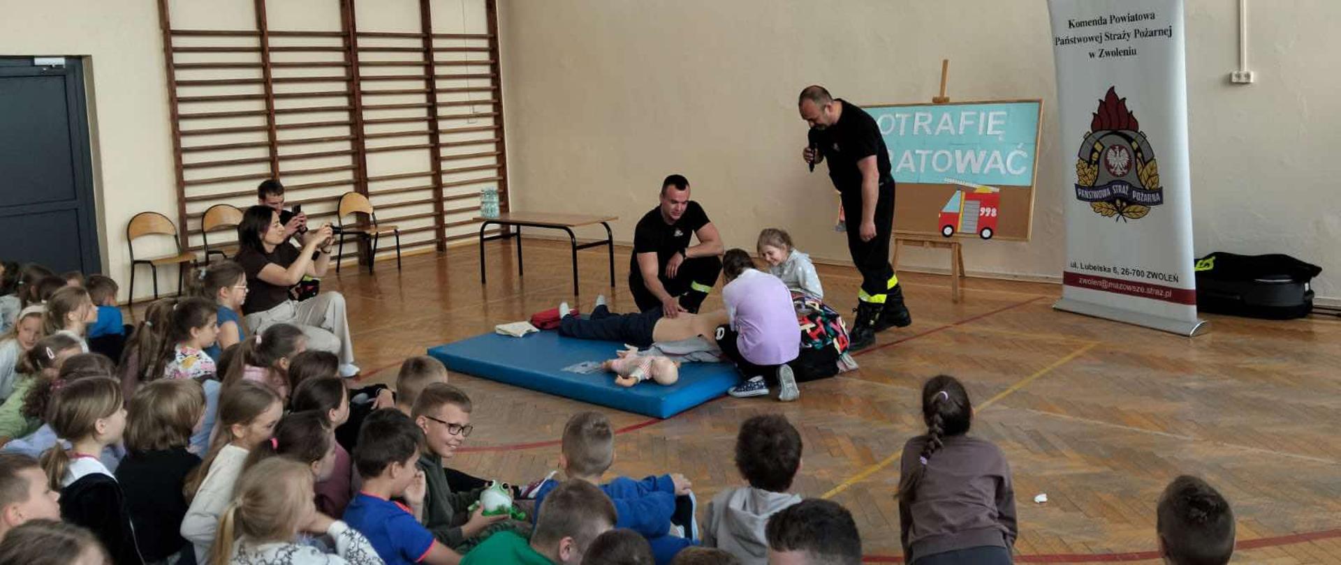 Zdjęcie przedstawia pokaz pierwszej pomocy prowadzony przez strażaków dla grupy dzieci, prawdopodobnie w sali gimnastycznej. Dwóch strażaków, jeden z mikrofonem, demonstruje resuscytację na manekinie leżącym na niebieskiej macie. Dzieci siedzą na podłodze, obserwując pokaz. W tle widoczny jest baner Komendy Powiatowej Państwowej Straży Pożarnej w Zwoleniu z hasłem "POTRAFIĘ RATOWAĆ" i grafiką wozu strażackiego. Sala jest jasna, z drewnianą podłogą i drabinkami na ścianie.