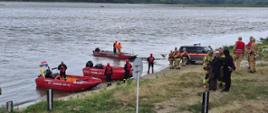 Strażacy z łodziami i samochodem stoją na brzegiem Wisły porośniętym trawą. Część z nich stoi w łodziach z zapiętymi kamizelkami ratowniczymi. 