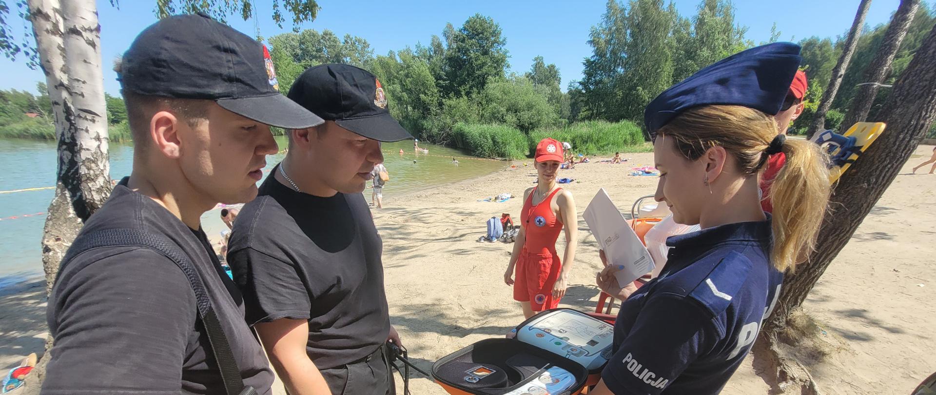 Na zdjęciu widoczni są funkcjonariusze straży pożarnej oraz policjantka trzymająca w rękach AED. W tle widać ratownika WOPR który opowiada o urządzeniu. Dalej widoczni są kąpiący się w stawie ludzie. Jest piękna, słoneczna pogoda. 
