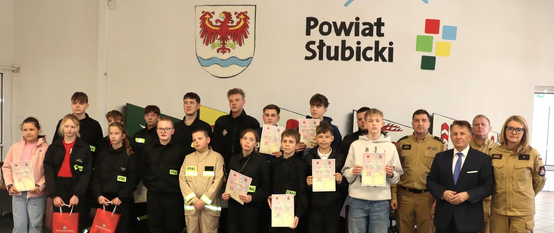 Komendant Powiatowy PSP, starosta i dzieci biorące udział konkursie OTWP pozują do grupowego zdjęcia. Dzieci trzymają torby z nagrodami oraz dyplomy.