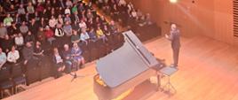 Widok z góry, artysta stoi przy fortepianie i mówi do mikrofonu, w tle widać widownię z publicznością
