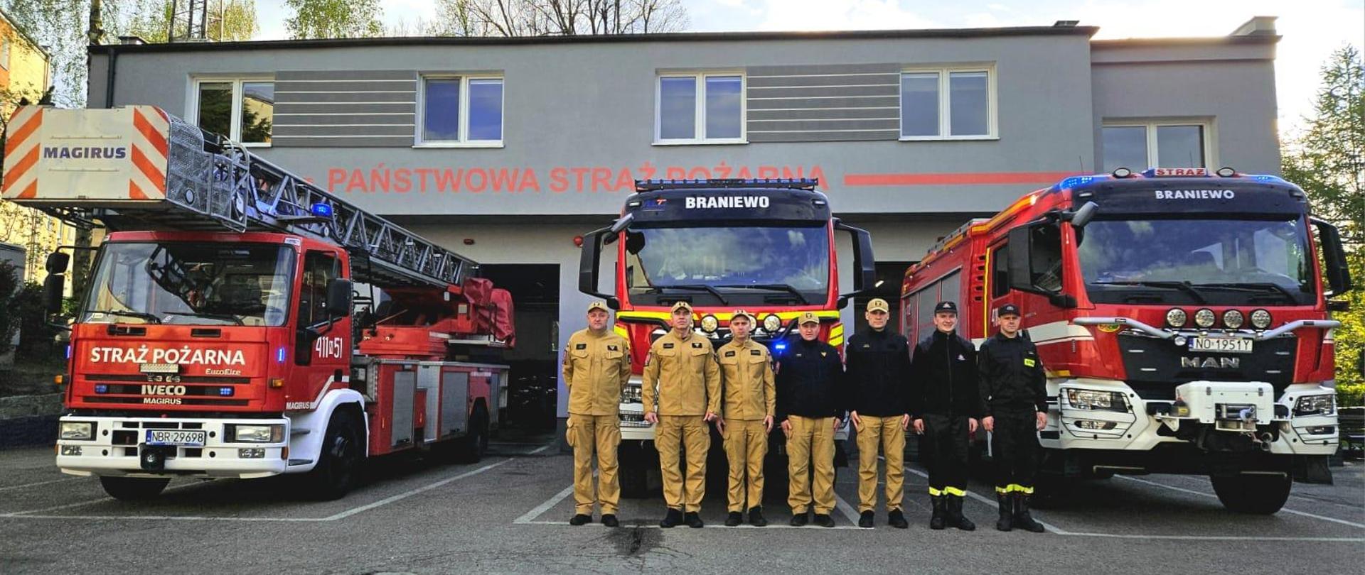 Strażacy w mundurach na tle samochodów strażackich oraz remizy.