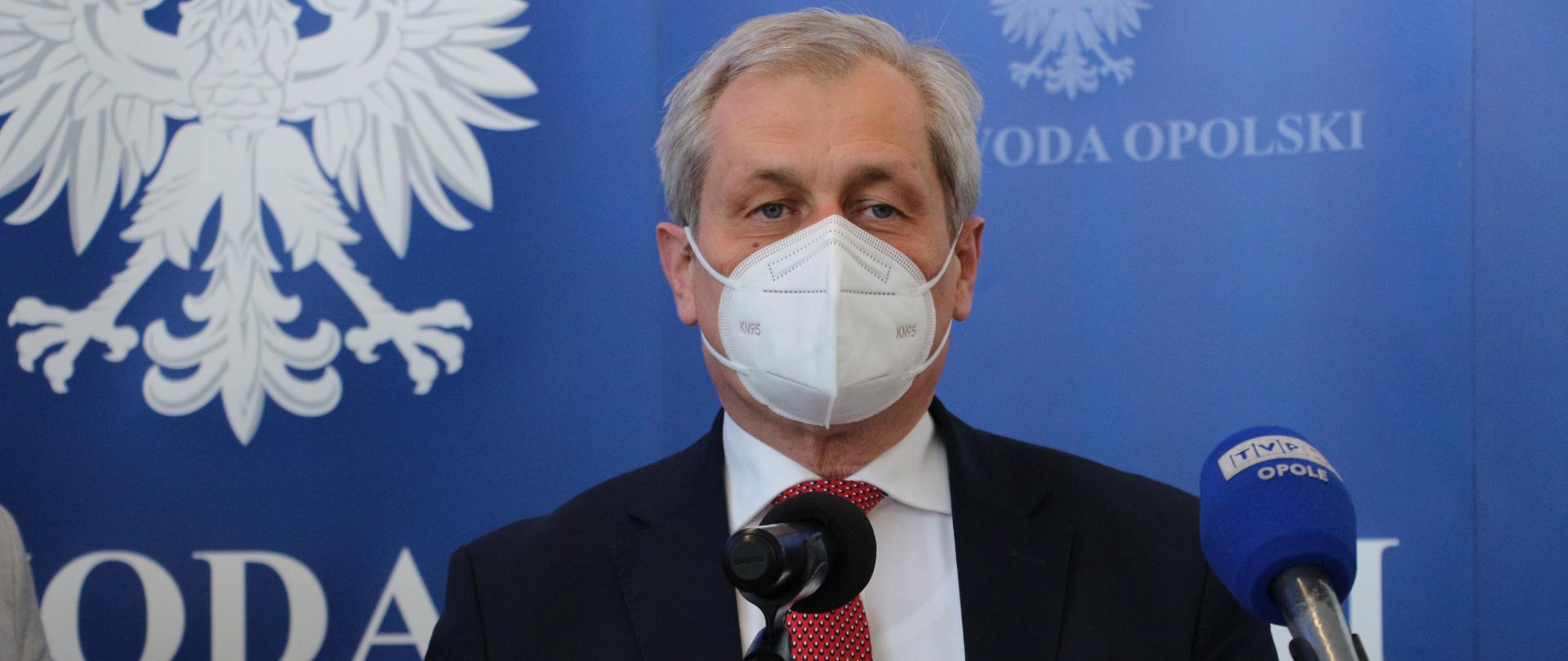 Na zdjęciu wojewoda opolski Sławomir Kłosowski, ubrany w czarny garnitur, białą koszulę i czerwony krawat we wzroki. Stoi przed szklaną mównicą, która sięga mu na wysokość klatki piersiowej. Przed mównicą stoją dwa statywy z mikrofonami. Wojewoda przemawia do zgromadzonych na konferencji prasowej. W tle ścianka granatowa z napisem Wojewoda Opolski. 