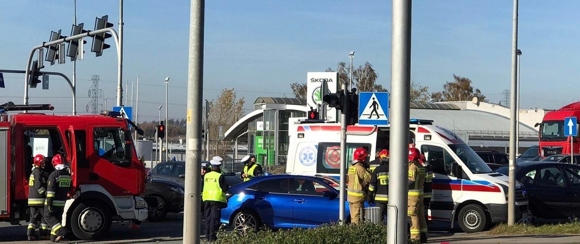 1. Niebieski samochód stoi na środku skrzyżowania. Przy nim stoją strażacy w umundurowaniu w kolorze piaskowym, za nimi biała karetka pogotowia. Po lewej stronie czerwony wóz strażacki a przy nim strażacy w mundurach bojowym w kolorze granatowym