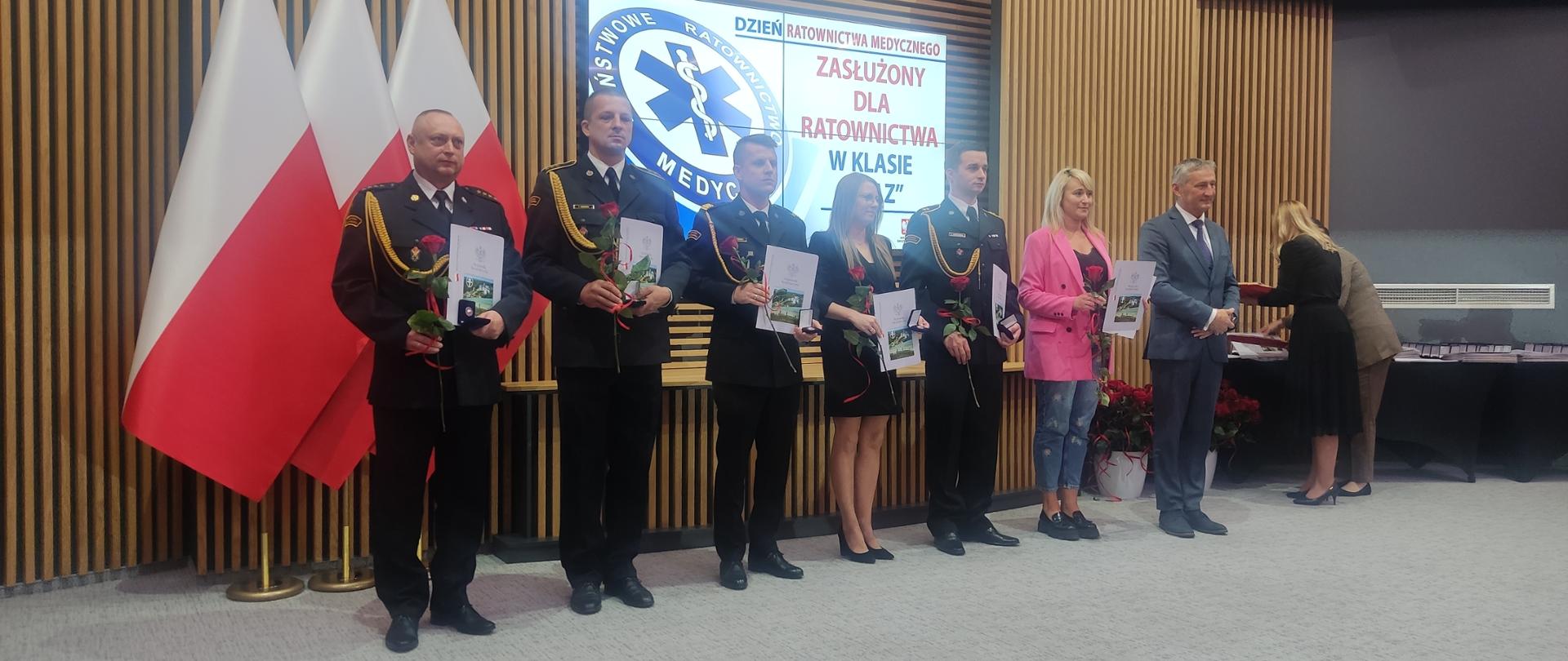 Na zdjęciu widzimy 4 strażaków w szeregu po odebraniu odznaczeń. Oprócz strażaków w szeregu stoją też trzy osoby cywilne w odświętnych ubraniach.