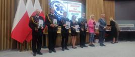 Na zdjęciu widzimy 4 strażaków w szeregu po odebraniu odznaczeń. Oprócz strażaków w szeregu stoją też trzy osoby cywilne w odświętnych ubraniach.
