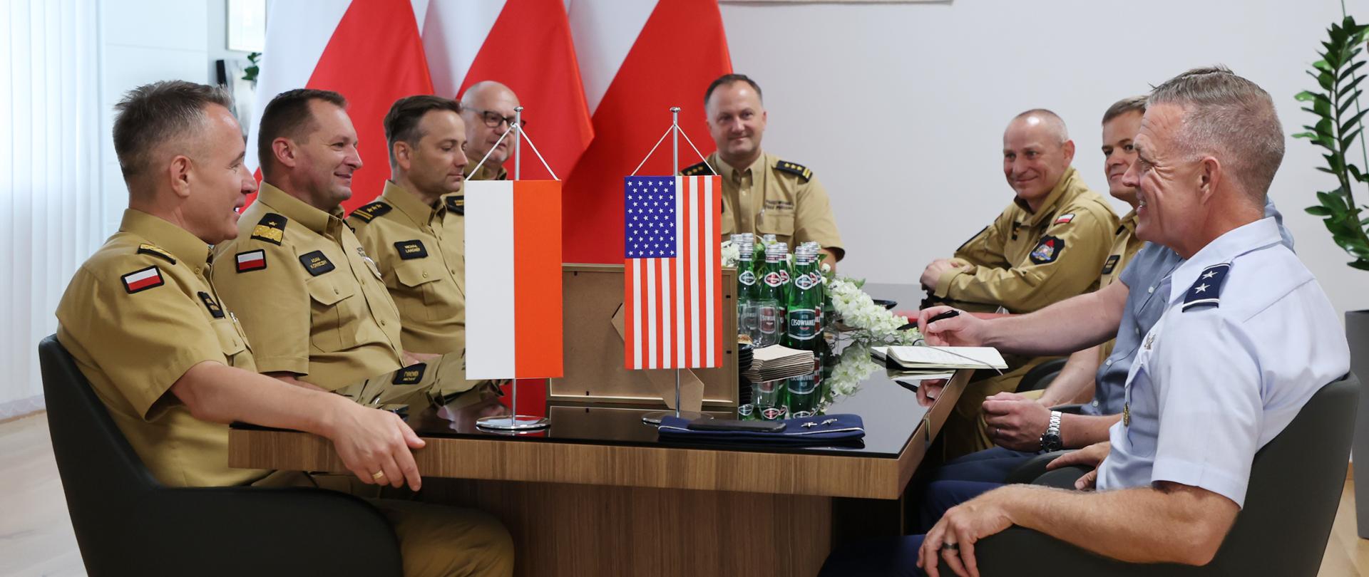 Gabinet komendanta głównego. Spotkanie kierownictwa komendy głównej z przedstawicielkami amerykańskiej gwardii narodowej. Na zdjęciu na pierwszym planie widoczny jest stół konferencyjny przy którym siedzą: komendant główny z zastępcą, dyrektorzy biur oraz dwuosobowa amerykańska delegacja. Na stole widoczne są proporce z flagami. W tle na ścianie widoczne godło i trzy polskie flagi. 