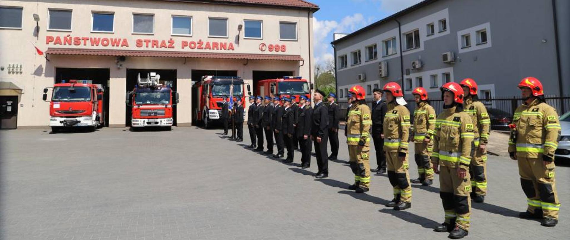 Zdjęcie przedstawia funkcjonariuszy Komendy Powiatowej Państwowej Straży Pożarnej w Zambrowie ustawionych na placu przed Komendą Powiatową Państwowej Straży Pożarnej w dwuszeregu. Część funkcjonariuszy ubrana w ubranie bojowe, pozostała część w mundury wyjściowe ze sznurem galowym. W tle widać front budynku Komendy Powiatowej Państwowej Straży Pożarnej w Zambrowie oraz pojazdy stanowiące wyposażenie Jednostki Ratowniczo-Gaśniczej w Zambrowie. 