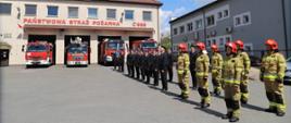 Zdjęcie przedstawia funkcjonariuszy Komendy Powiatowej Państwowej Straży Pożarnej w Zambrowie ustawionych na placu przed Komendą Powiatową Państwowej Straży Pożarnej w dwuszeregu. Część funkcjonariuszy ubrana w ubranie bojowe, pozostała część w mundury wyjściowe ze sznurem galowym. W tle widać front budynku Komendy Powiatowej Państwowej Straży Pożarnej w Zambrowie oraz pojazdy stanowiące wyposażenie Jednostki Ratowniczo-Gaśniczej w Zambrowie. 