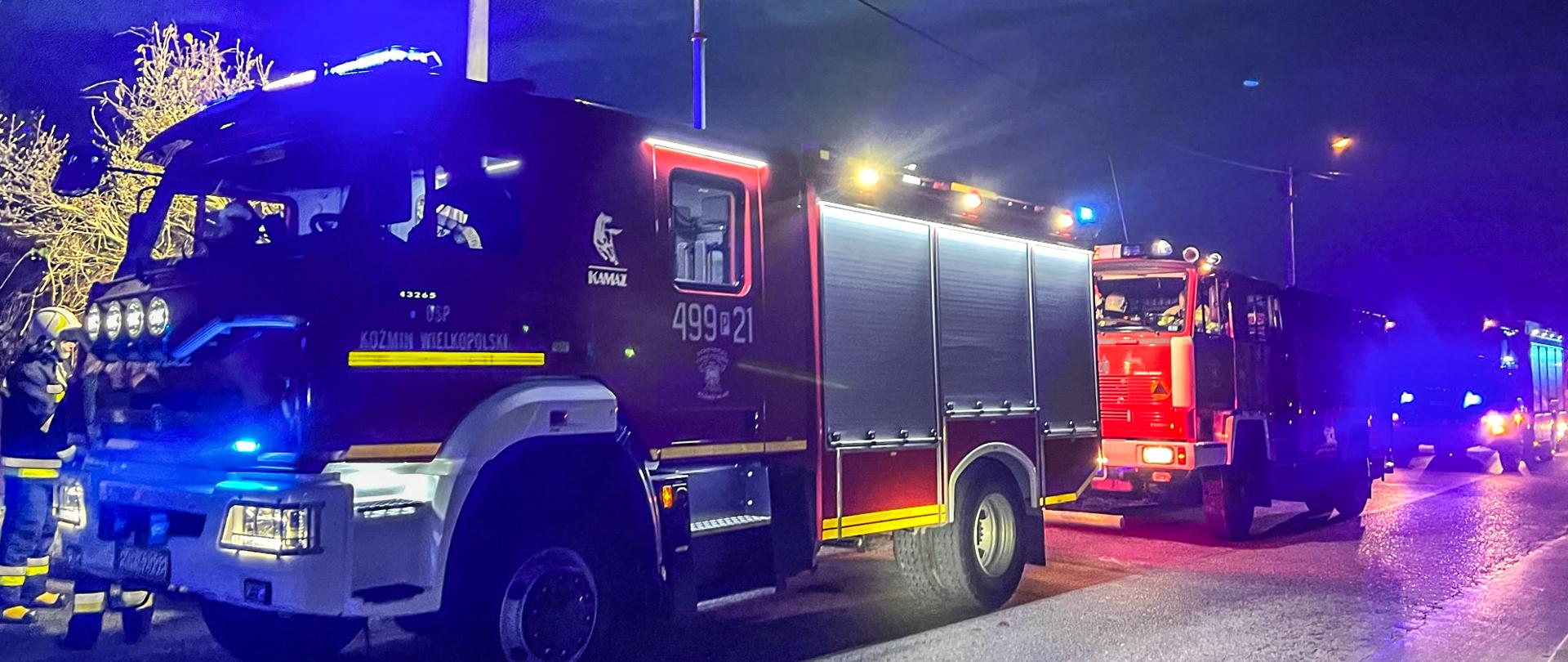 Na drodze przebiegającej wzdłuż miejsca prowadzonych działań znajdują się pojazdy pożarnicze ustawione w jednym ciągu. W pojazdach włączone jest pełne oświetlenie oraz sygnalizacja świetlna. Na pierwszym planie widoczny pojazd pożarniczy jednostki OSP Koźmin, który dodatkowo realizuje oświetlenie terenu działań za pomocą masztu oświetleniowego. Obok pojazdów kręcą się strażacy. Zdjęcie wykonane w porze nocnej.