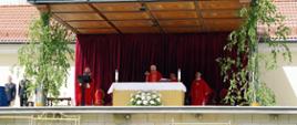 Widok z przodu. Celebrans w trakcie odprawiania liturgii na ołtarzu polowym. Z ambony zlokalizowanej po lewej stronie przybyłych na uroczystość wita kustosz sanktuarium. Z jego lewej strony strażacy pełniący funkcję służby liturgicznej. 