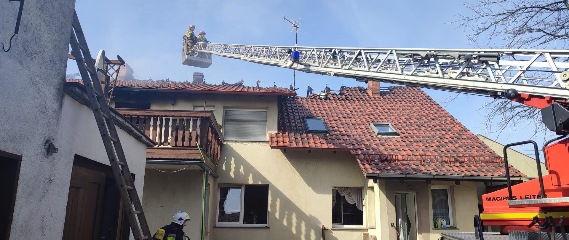 Na zdjęciu znajduje się budynek z nadpalonym dachem oraz drabina pożarnicza ze strażakami w koszu