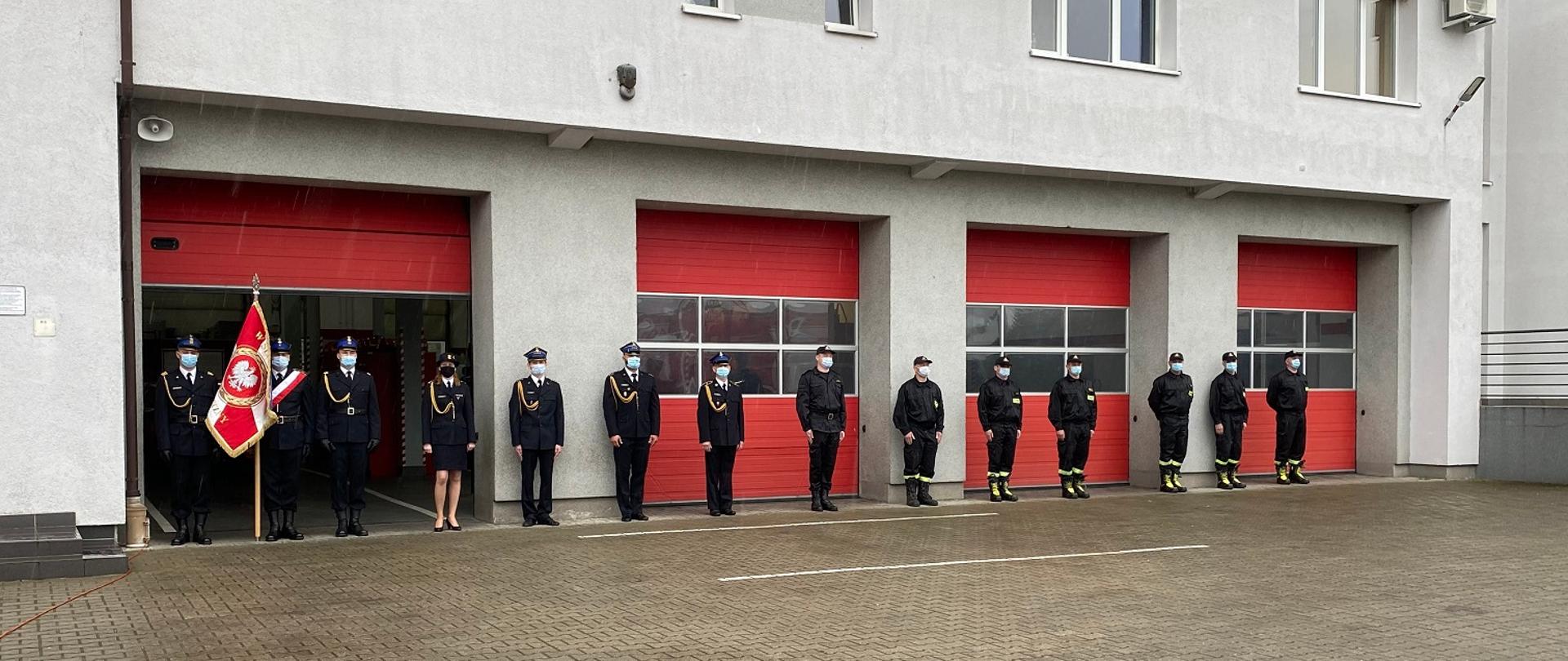 Funkcjonariusze PSP ustawieni w szeregu. W tle widoczny budynek straźnicy.