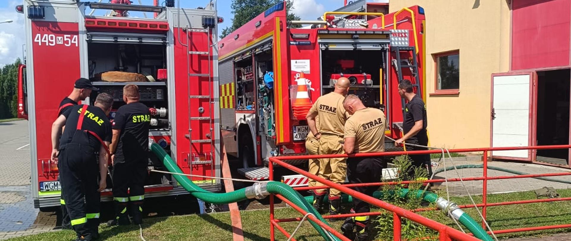 Zdjęcie przedstawia dwóch strażaków podłączających zielony, gruby wąż do czerwonego wozu strażackiego z oznaczeniem "449 54". Wokół wozów strażackich znajdują się inni strażacy, niektórzy w ciemnych ubraniach koszarowych, inni w jasnych ubraniach koszarowych, przygotowujący sprzęt. W tle widać budynek straży pożarnej z czerwonymi drzwiami garażowymi oraz maszt antenowy. Teren wokół wozów jest utwardzony kostką brukową.