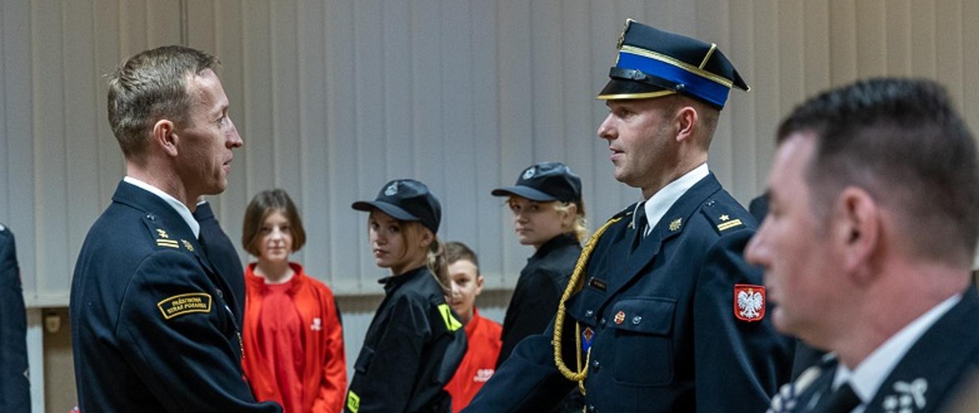 strażak w mundurze wita się z innym strażakiem w mundurze w tle dzieci