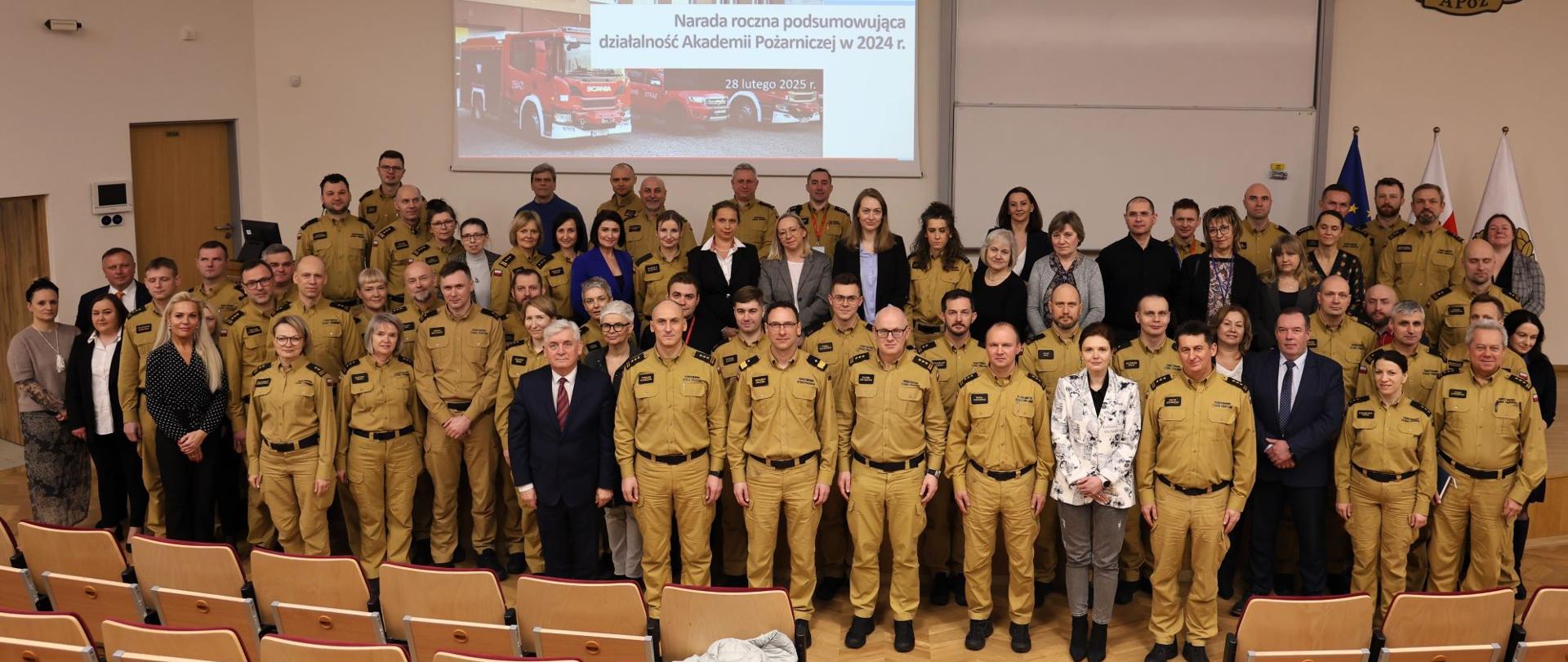 Zdjęcie grupowe uczestników konferencji w audytorium. Na tle widoczny jest ekran z prezentacją zatytułowaną „Narada roczna podsumowująca działalność Akademii Pożarniczej w 2024 r.”. Większość uczestników ubrana jest w mundury, inni w stroje formalne. Zdjęcie wykonane jest z perspektywy audytorium, pokazując pełną salę