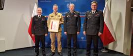 Strażacy w mundurach stoją w szeregu jeden trzyma dyplom na tle flag polski.