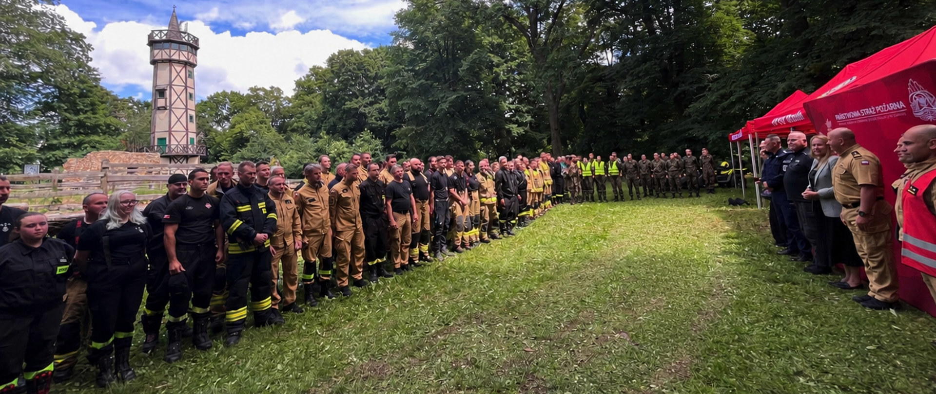 Grupa kilkudziesięciu strażaków, ratowników i służb mundurowych stoi w szeregu podczas uroczystej zbiórki na polanie otoczonej wysokimi drzewami. Po prawej stronie znajdują się czerwone namioty Państwowej Straży Pożarnej, przed którymi stoją przedstawiciele organizatorów i goście. W tle widoczna jest wieża obserwacyjna. Nad uczestnikami rozciąga się błękitne niebo z białymi chmurami. Zdjęcie wykonano w słoneczny dzień podczas wydarzenia lub ćwiczeń służb ratowniczych.