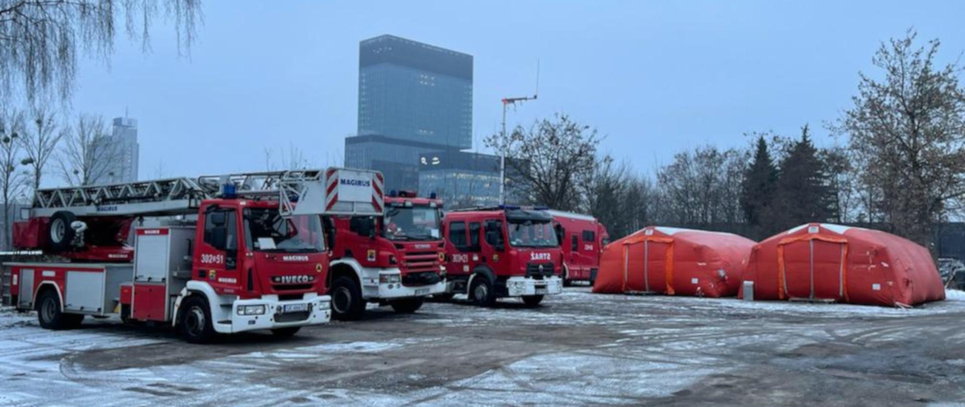 Na zdjęciu widoczny jest plac, na którym po lewej stronie stoją zaparkowane dwa samochody gaśnicze i samochód specjalny drabina oraz samochód dowodzenia i łączności. Na wprost widoczne są pomarańczowe namioty pneumatyczne Państwowej Straży Pożarnej. Na dalszym planie widoczny jest szklany budynek biurowy i Międzynarodowego Centrum Kongresowego.
